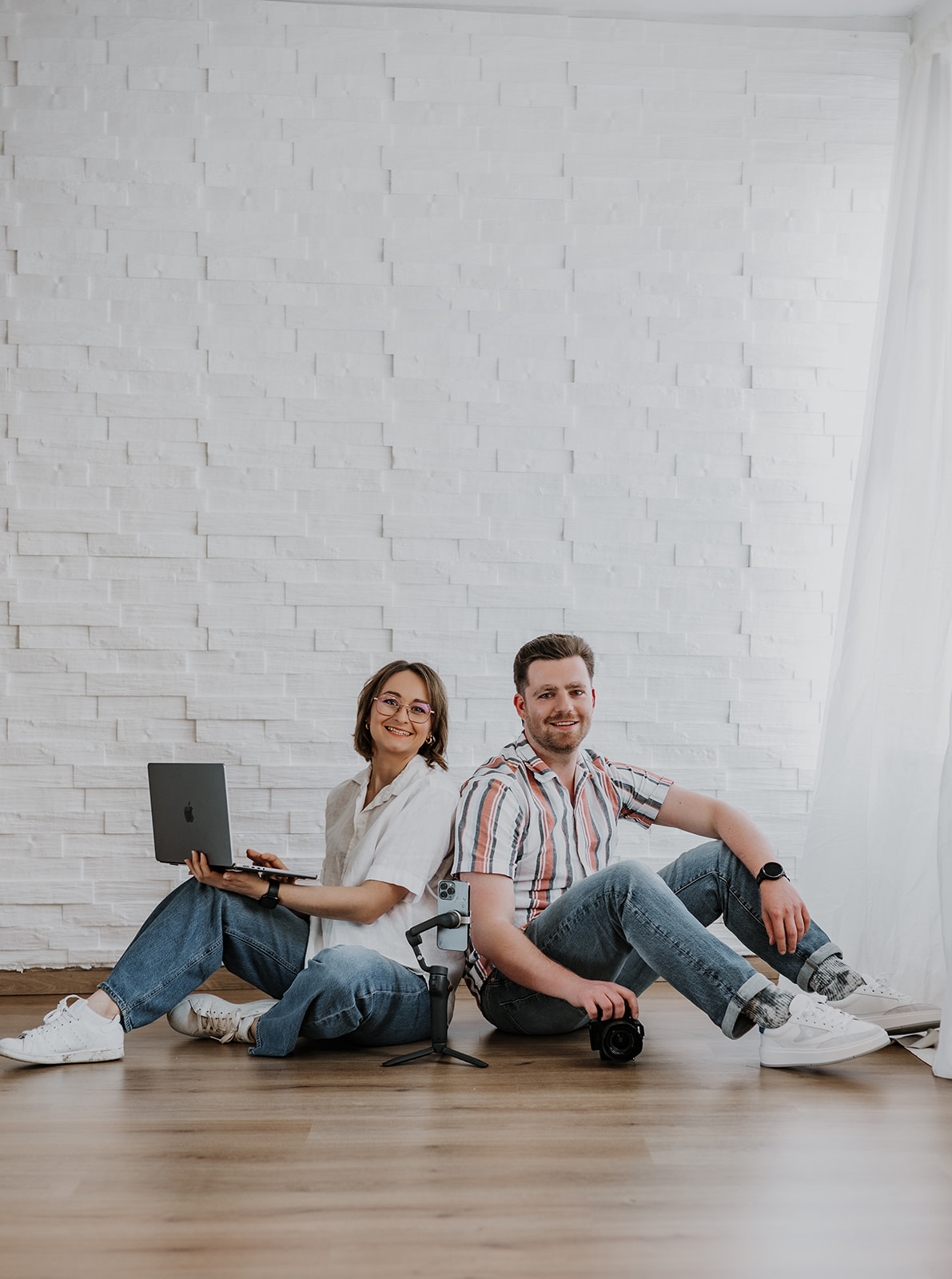 Die Geschäftsführer sitzen vor einer weißen Wand auf dem Boden mit Equipment und blicken in die Kamera.